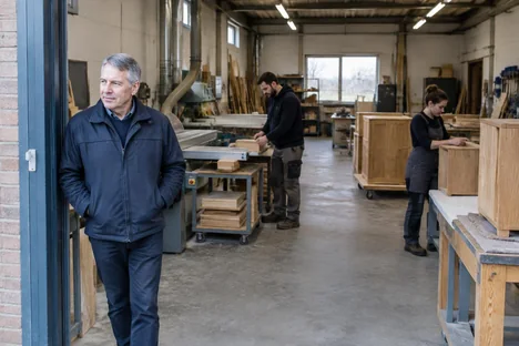 An experienced man at a furniture workshop doorway while colleagues continue working
